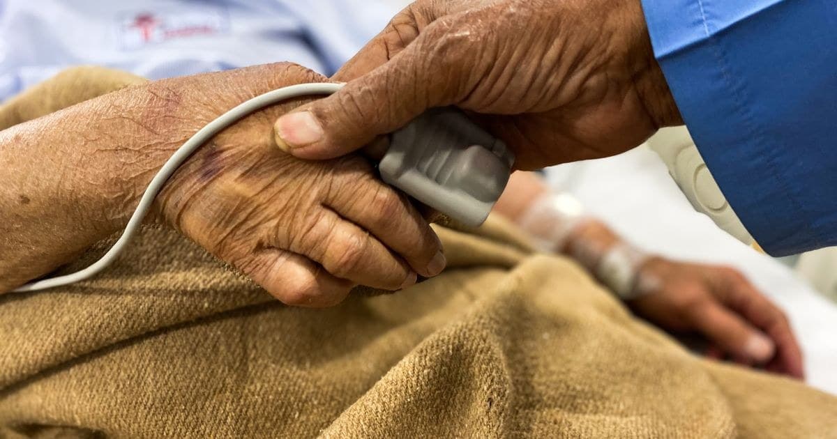 Mano de una paciente mayor con un oxímetro en el dedo, sostenida por la mano de un familiar en una cama de hospital.